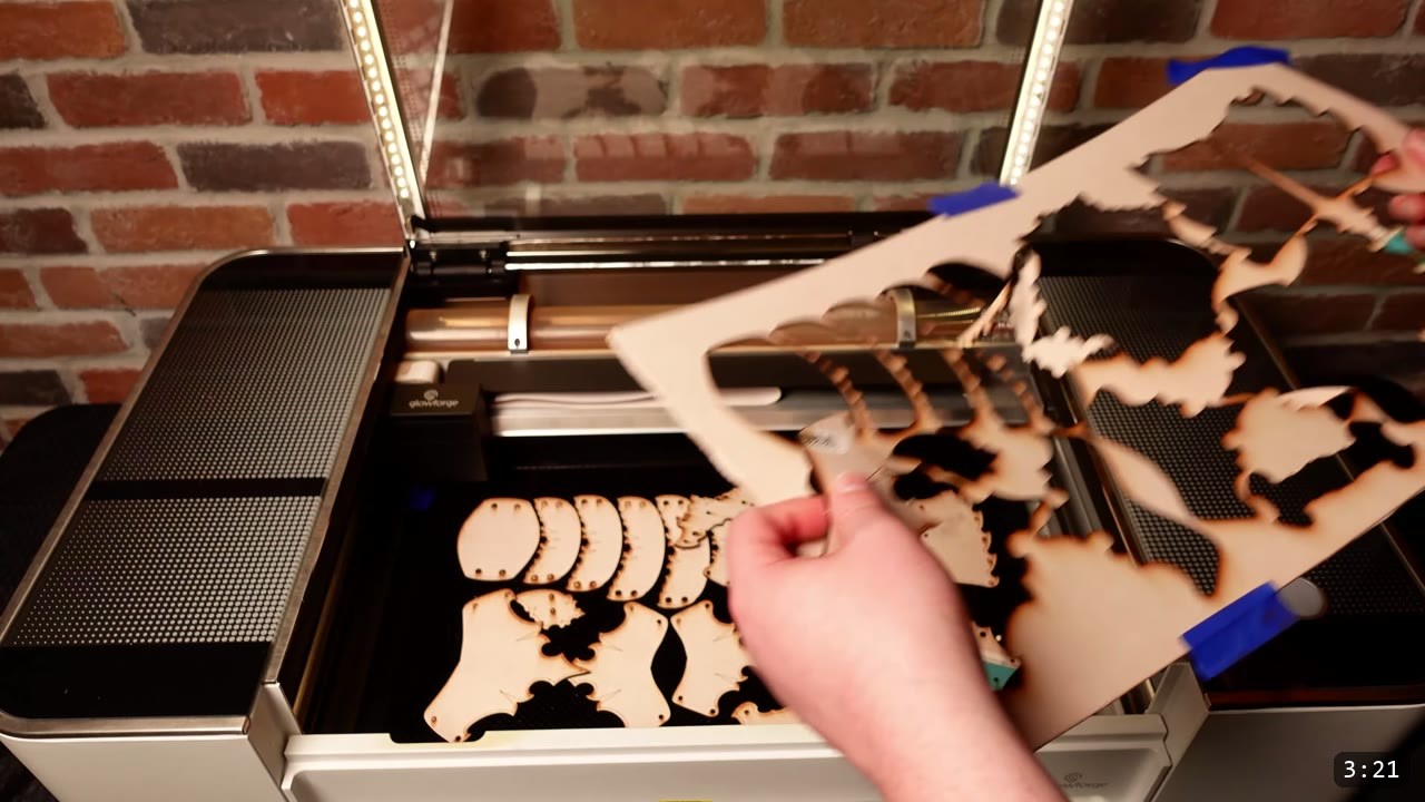 Articulating Leather Gauntlets Build Guide: Imperial Knight, Cut to Finish 2 a Glowforge laser cutter open with a hand lifting the cut natural-leather waste sheet away, small laser-cut plates and scales left in the bed, brick wall behind