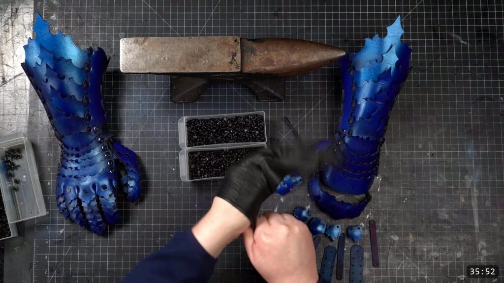 both finished blue articulating leather gauntlets standing upright with full jointed finger scales, a horned anvil and two trays of black rivets between them