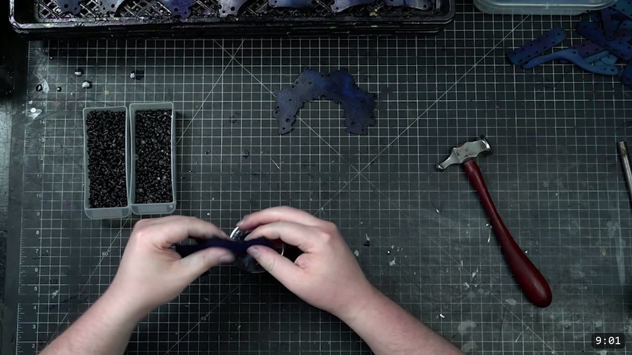 Articulating Leather Gauntlets Build Guide: Imperial Knight, Cut to Finish 5 a top-down cutting mat with two hands folding a dyed dark-blue knuckle plate over a rivet, trays of black rivets and a small hammer nearby
