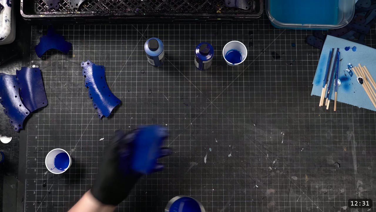 Articulating Leather Gauntlets Build Guide: Imperial Knight, Cut to Finish 6 a top-down cutting mat with glossy blue-painted plates, small paint bottles and a cup of blue paint, a black-gloved hand holding a blue piece, and blue-stained stir sticks on a foam block
