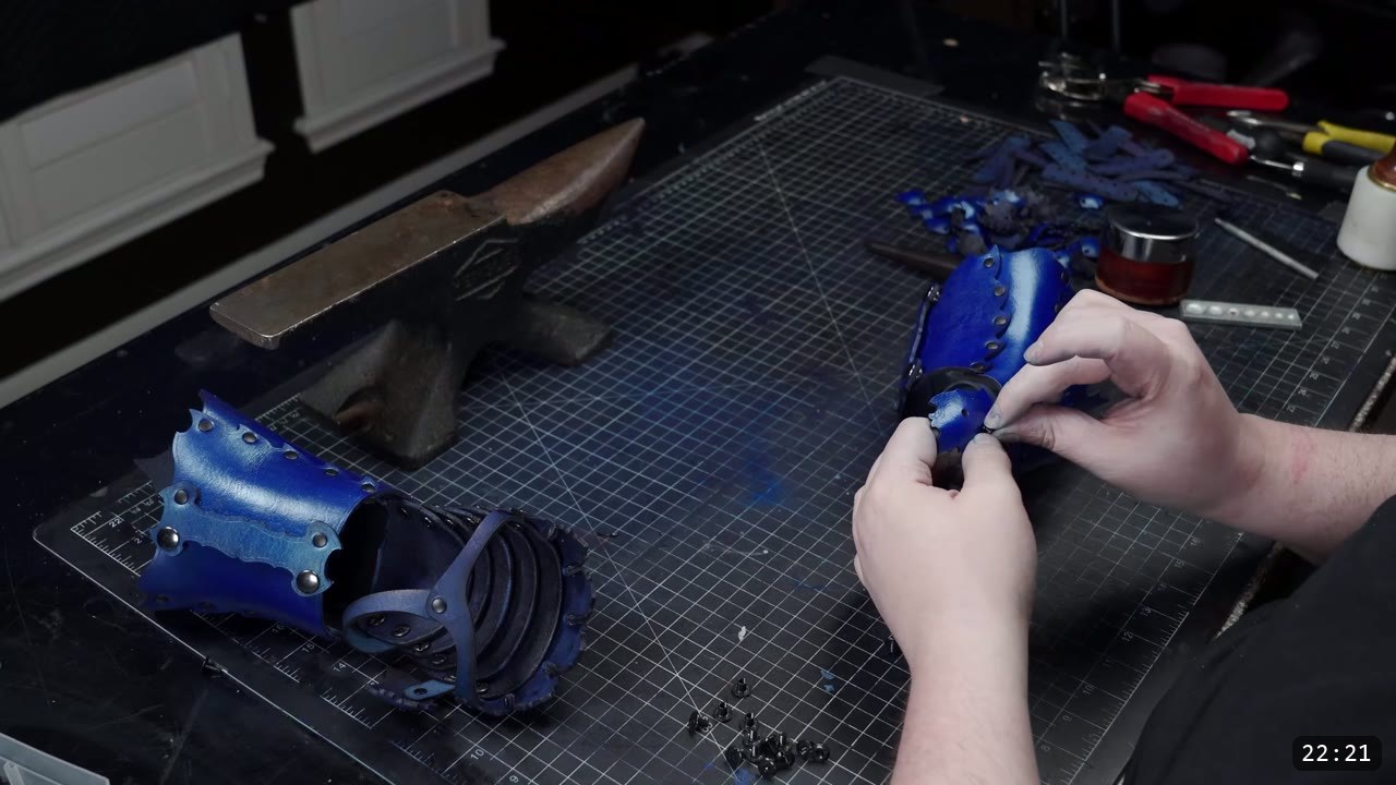 Articulating Leather Gauntlets Build Guide: Imperial Knight, Cut to Finish 8 a bench with a finished blue wrist cuff beside an anvil, two hands riveting a blue wrist piece, scattered black rivets and pliers on the mat