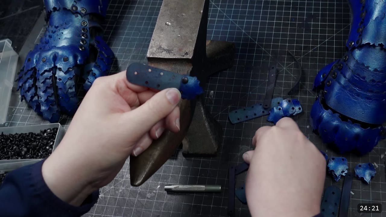 Articulating Leather Gauntlets Build Guide: Imperial Knight, Cut to Finish 9 two blue articulated gauntlets standing at left and right with a pair of hands setting a rivet on a small finger plate against the anvil horn, a tray of black rivets and a center punch nearby