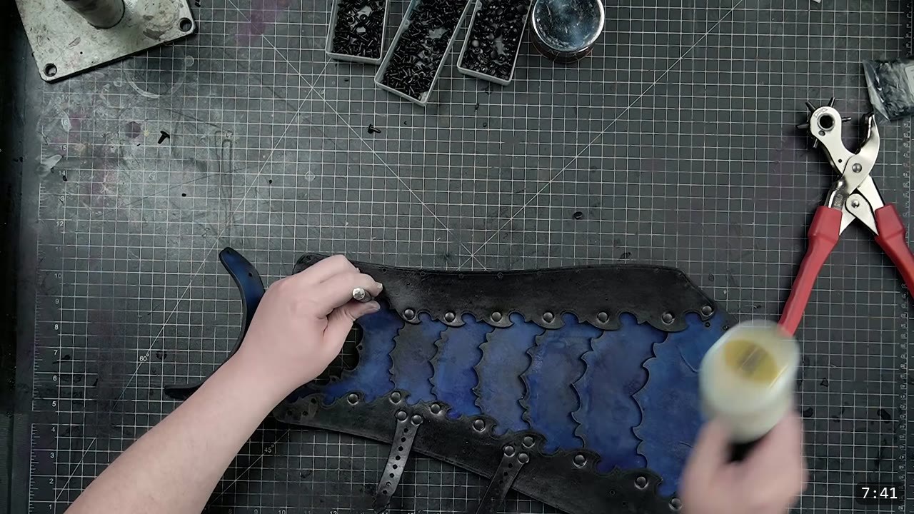 Leather Greaves Build Guide: Imperial Leg Armor from a Flat Pattern 9 overhead view of the assembled flat greave back section, black with riveted blue lamellar scallops, a rotary punch and finish bottle beside it