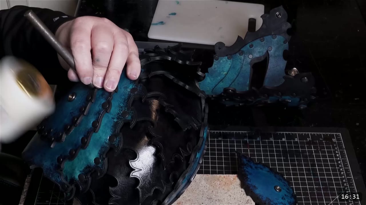 a hand setting rivets with a mallet and setter on the assembled dark-dyed helmet, with a matching faceplate piece nearby