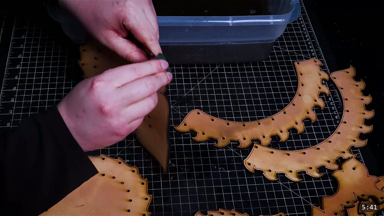 a green scrubbing pad cleaning the edges of a damp tan leather helmet piece on a gridded cutting mat, with other punched pieces nearby