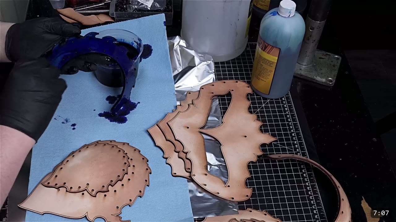 black-gloved hands mixing dark blue-black dye in a tray beside tan leather helmet panels, with a bottle of dye on the bench
