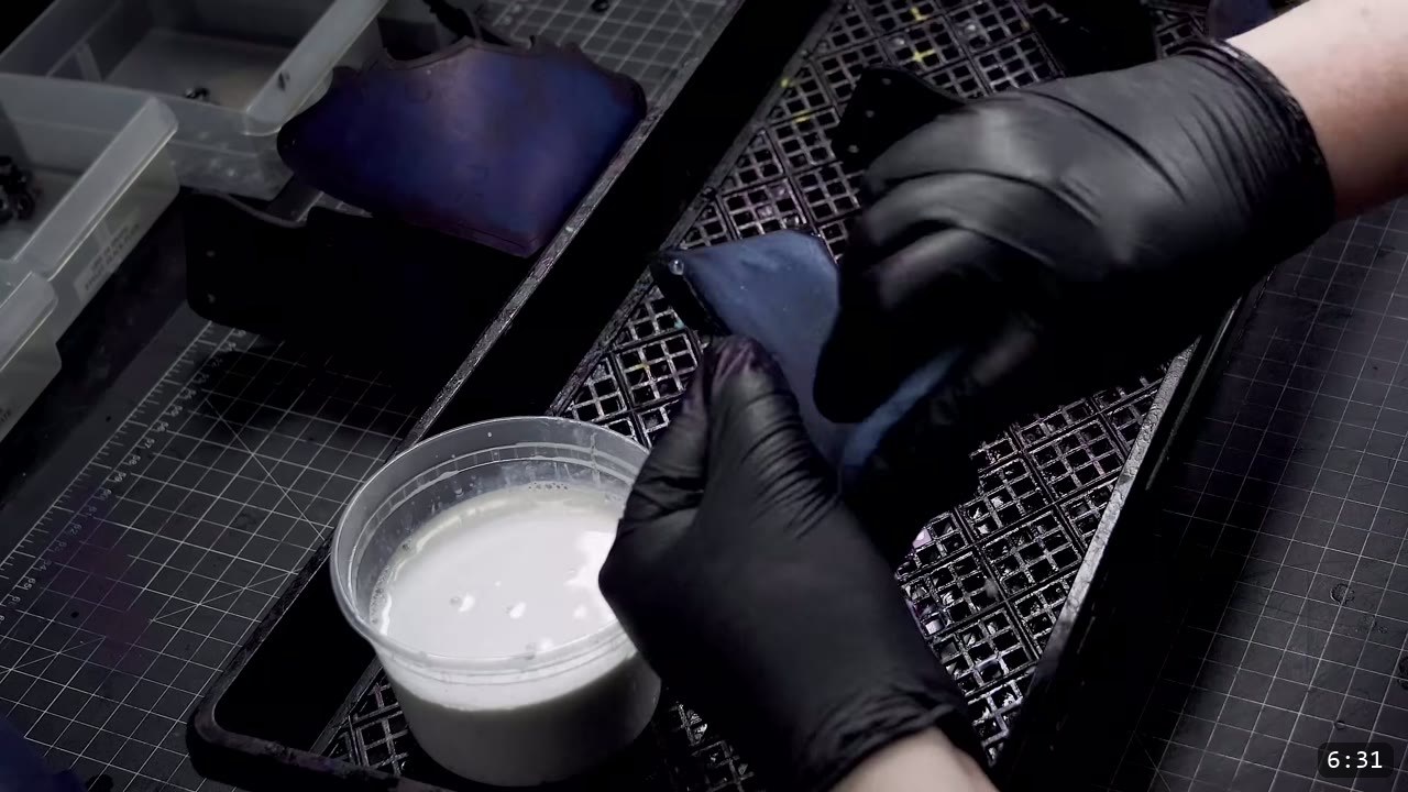 gloved hands working a dyed deep-blue piece over a rack beside a tub of light-colored finishing liquid