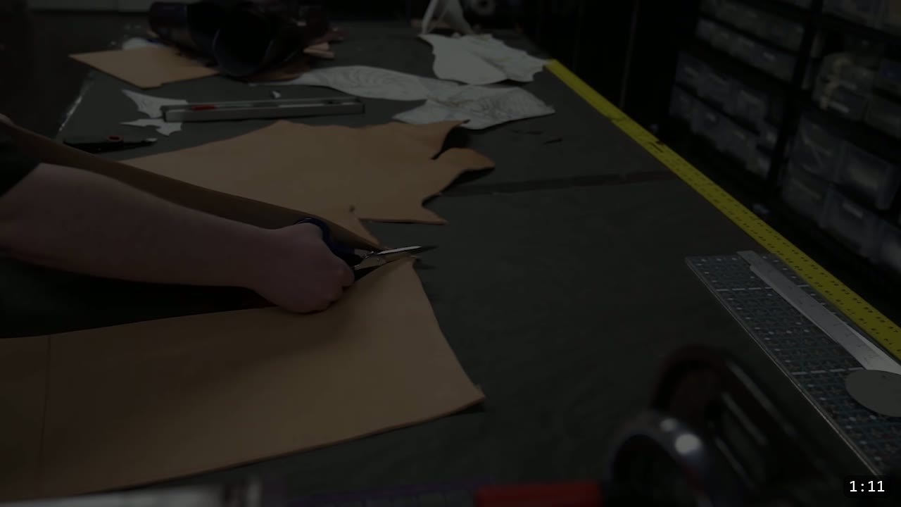 Imperial Knight Tassets Build Guide: Leather Hip Armor from a Flat Pattern 3 hands cutting a large piece of tan vegetable-tan leather with shears on a dark workbench, paper pattern pieces and a straight edge nearby