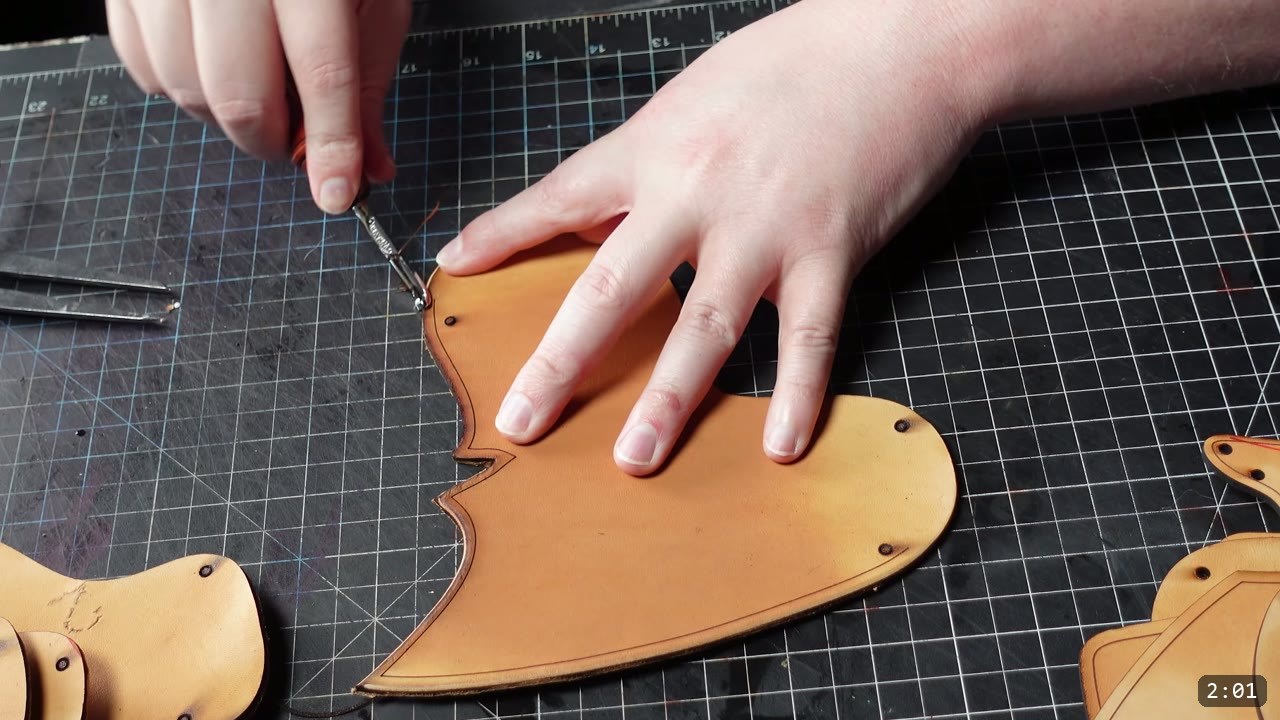 Imperial Knight Tassets Build Guide: Leather Hip Armor from a Flat Pattern 5 hands running a small edge tool along the top edge of a cut tan tasset piece on a grid cutting mat, other cut plates with punched holes nearby