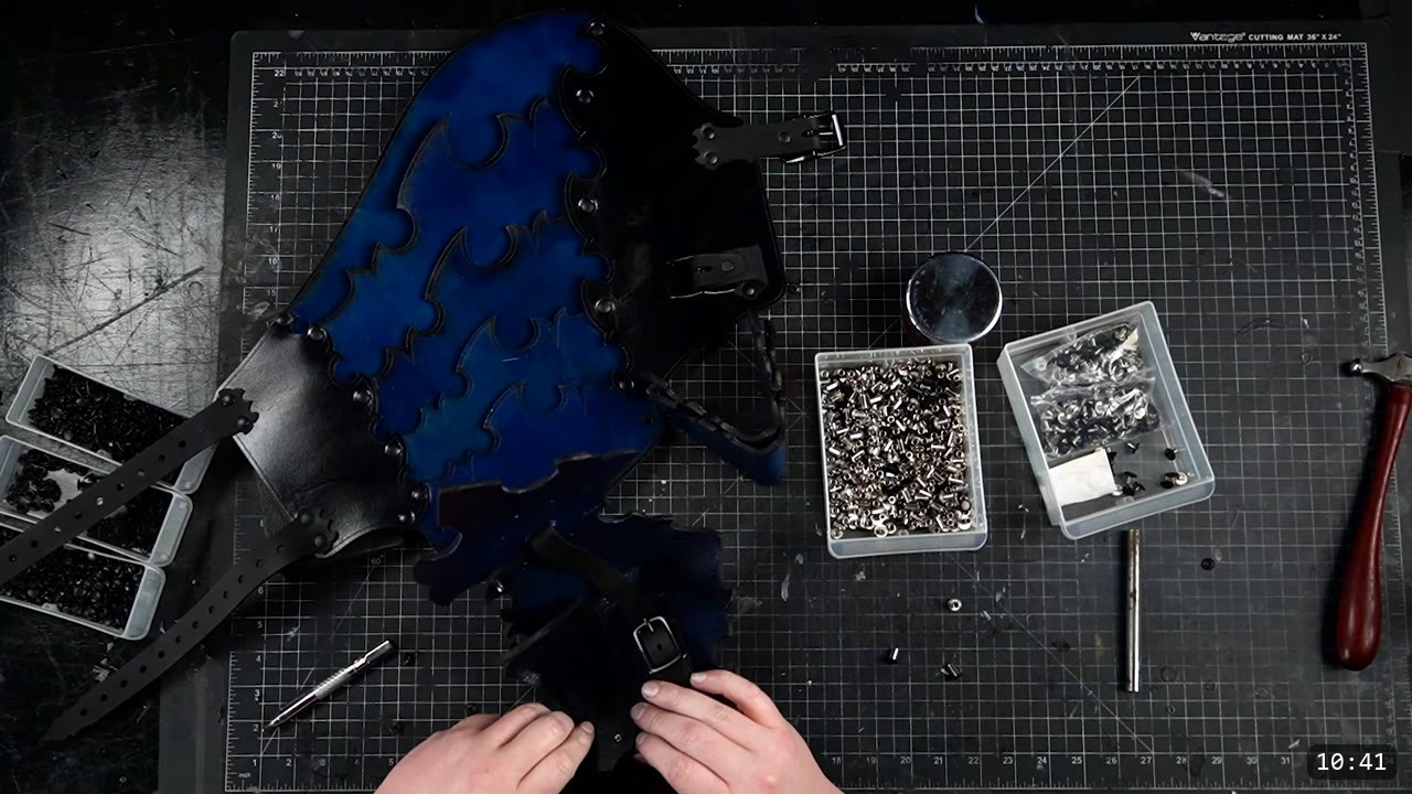 Leather Leg Armor Build Guide: Articulated Cuisses, Knees, and Greaves 12 hands attaching a strap at the base of a royal-blue leg plate, two open bins of silver Chicago screw posts and black hardware on the bench