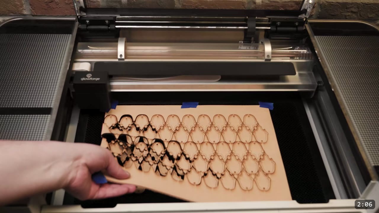 Leather Scale Skirting Build Guide: Laser-Cut Scales into a Modular Armor Skirt 2 a Glowforge laser cutter with a tan leather sheet taped down with blue tape and a full field of cut scale outlines, some edges scorched, a hand lifting the sheet