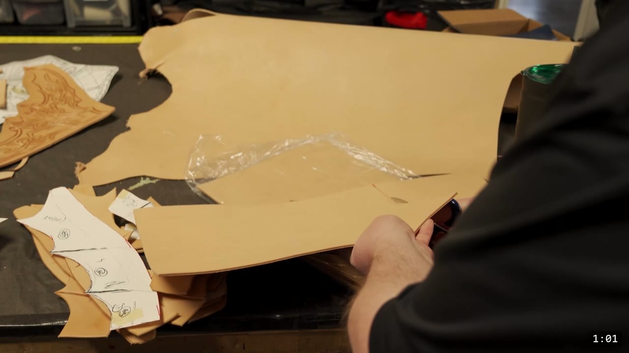 Leather Scale Skirting Build Guide: Laser-Cut Scales into a Modular Armor Skirt 4 a large tan vegetable-tan leather piece on a dark workbench with paper pattern templates and a tooled scrap to the left and a hand holding a phone