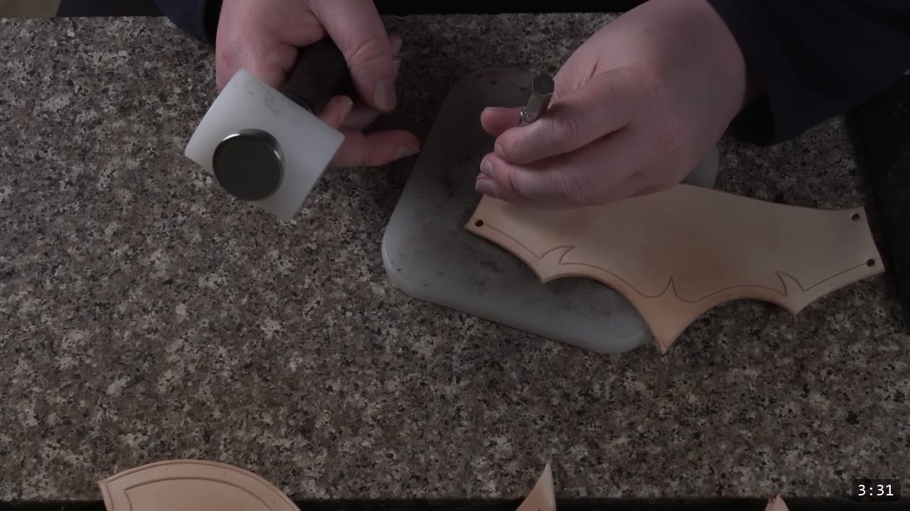 Leather Armor for Beginners: Tools You Need and How to Start 7 hands holding a white poly-head mallet and a hole punch over a green poundo board on a granite counter, punching a curved leather piece
