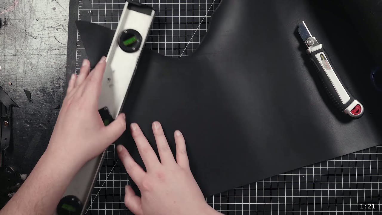 Leather Buckle Straps for Armor: A Reusable How-To with Free Patterns 4 using an aluminum level as a straight edge on a piece of black leather, a box cutter resting on the mat