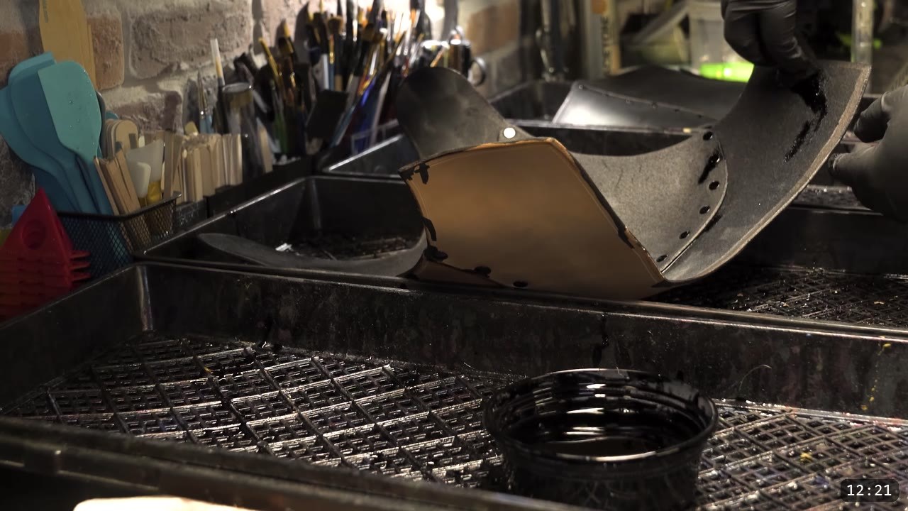 a leather plate half-dyed black at the dye station, a cup of black dye and a rack of brushes, gloved hands holding a dyed piece