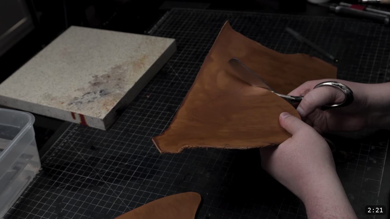 cutting a curved tan vegetable-tan breastplate panel with shears on a cutting mat, a granite slab nearby