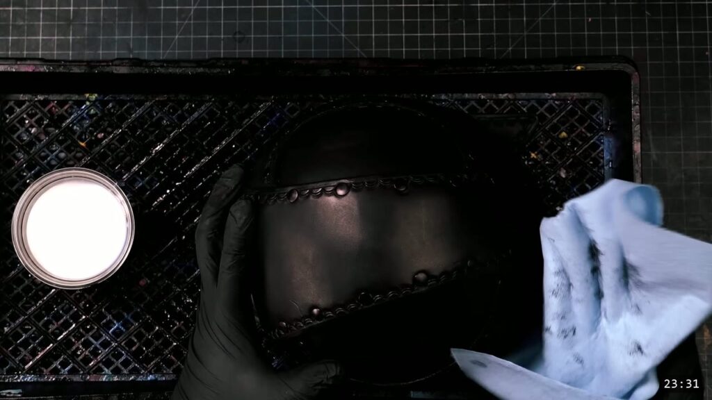 a black-gloved hand holding the dyed-black helmet over the dye rack, a tub of finish to the left, wiping the surface with a shop towel