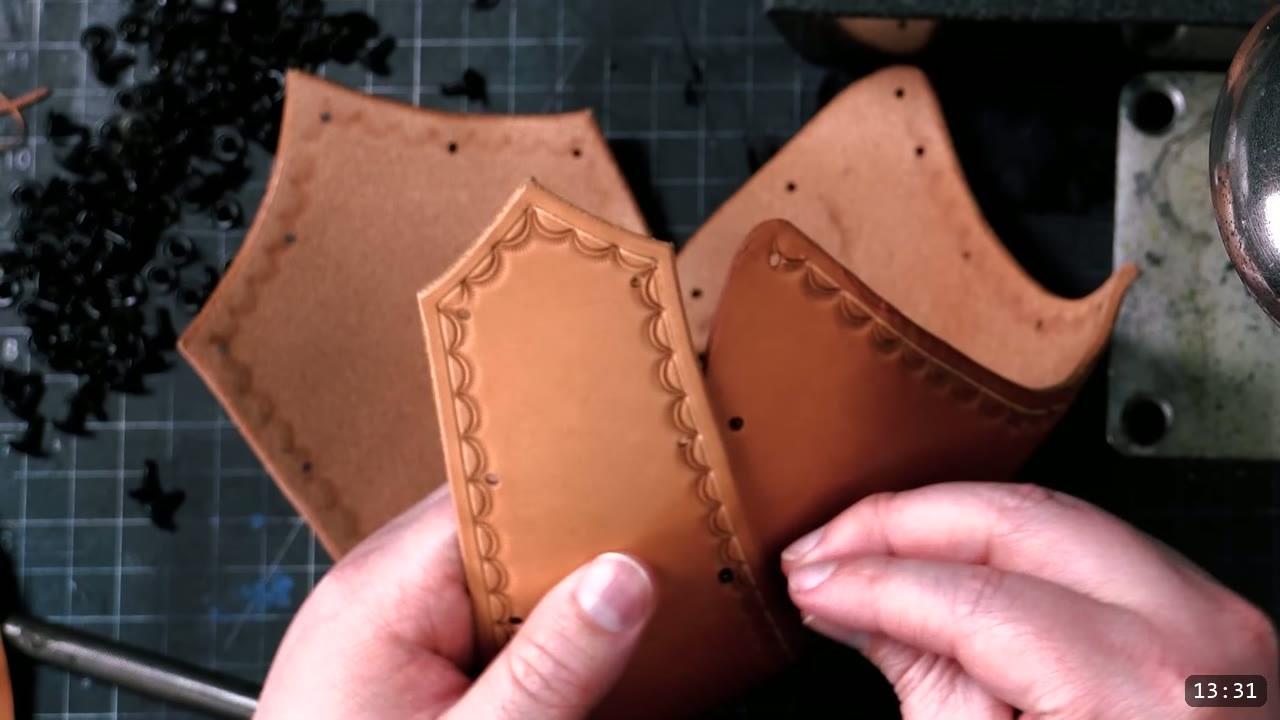 Leather Warrior Helmet Build Guide: Flat Pattern to Finished Piece 8 hands holding two tan helmet pieces with grooved and crescent-stamped borders, loose black double-cap rivets scattered on the mat